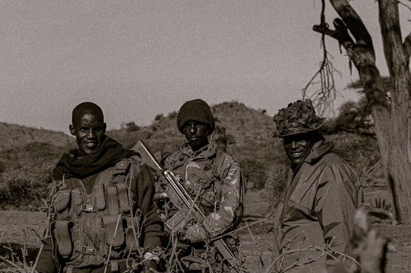 Three individuals stand outdoors, each wearing rugged clothing and gear typical of military or tactical environments. The setting is a natural landscape, possibly a rural or wilderness area, with trees and hills in the background. One person seems to be holding a firearm, and all appear dressed for outdoor or combat conditions.