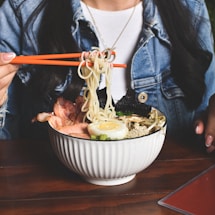 A person wearing a denim jacket and white shirt is holding orange chopsticks to lift noodles from a bowl of ramen, which is garnished with slices of pork, a halved boiled egg, seaweed, and green onions. The table surface is wooden and there's a glimpse of a red menu card.