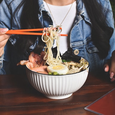 A person wearing a denim jacket and white shirt is holding orange chopsticks to lift noodles from a bowl of ramen, which is garnished with slices of pork, a halved boiled egg, seaweed, and green onions. The table surface is wooden and there's a glimpse of a red menu card.