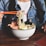 A person wearing a denim jacket and white shirt is holding orange chopsticks to lift noodles from a bowl of ramen, which is garnished with slices of pork, a halved boiled egg, seaweed, and green onions. The table surface is wooden and there's a glimpse of a red menu card.
