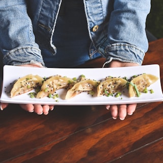 A person wearing a denim jacket holds a rectangular white plate with a row of dumplings arranged neatly. The dumplings are garnished with chopped green onions and black sesame seeds, sitting on a wooden table.