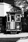 A small, enclosed booth with glass windows is situated in front of a brick building. The structure features steps leading up to a door and is placed on a patterned pavement. Shadows of trees and adjacent structures are cast on the building, creating a play of light and shadow.