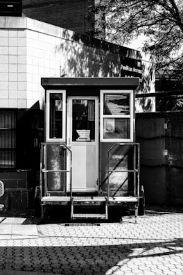 A small, enclosed booth with glass windows is situated in front of a brick building. The structure features steps leading up to a door and is placed on a patterned pavement. Shadows of trees and adjacent structures are cast on the building, creating a play of light and shadow.