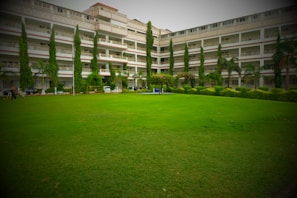A large, well-maintained lawn stretches out in front of a multi-story building. The building features balconies on each floor, with tall, slender trees and various plants lining the perimeter, providing a lush green landscape. A few people are visible near the building, and parked vehicles are partially visible behind the trees.