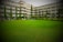 A large, well-maintained lawn stretches out in front of a multi-story building. The building features balconies on each floor, with tall, slender trees and various plants lining the perimeter, providing a lush green landscape. A few people are visible near the building, and parked vehicles are partially visible behind the trees.