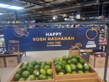 A supermarket display featuring a banner celebrating Rosh Hashanah is prominently visible. The sign includes festive graphics like a pomegranate and a honey jar, and text in both English and Hebrew. Below the sign, crates filled with green avocados are displayed. The overall setting suggests a promotion for a holiday event at a market.
