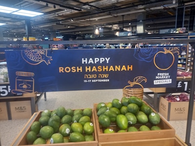 A supermarket display featuring a banner celebrating Rosh Hashanah is prominently visible. The sign includes festive graphics like a pomegranate and a honey jar, and text in both English and Hebrew. Below the sign, crates filled with green avocados are displayed. The overall setting suggests a promotion for a holiday event at a market.
