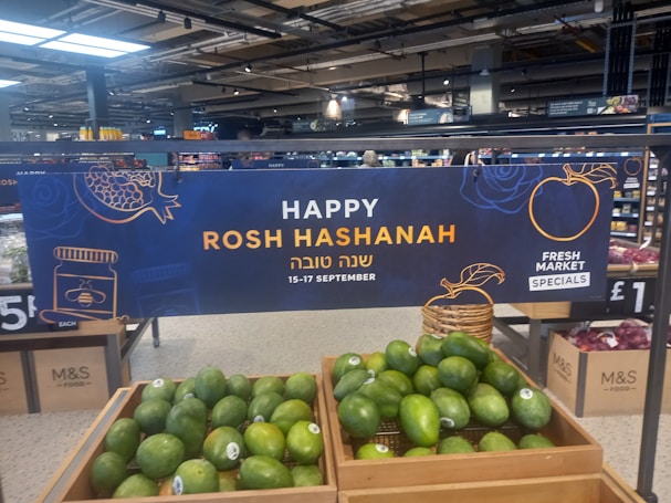 A supermarket display featuring a banner celebrating Rosh Hashanah is prominently visible. The sign includes festive graphics like a pomegranate and a honey jar, and text in both English and Hebrew. Below the sign, crates filled with green avocados are displayed. The overall setting suggests a promotion for a holiday event at a market.
