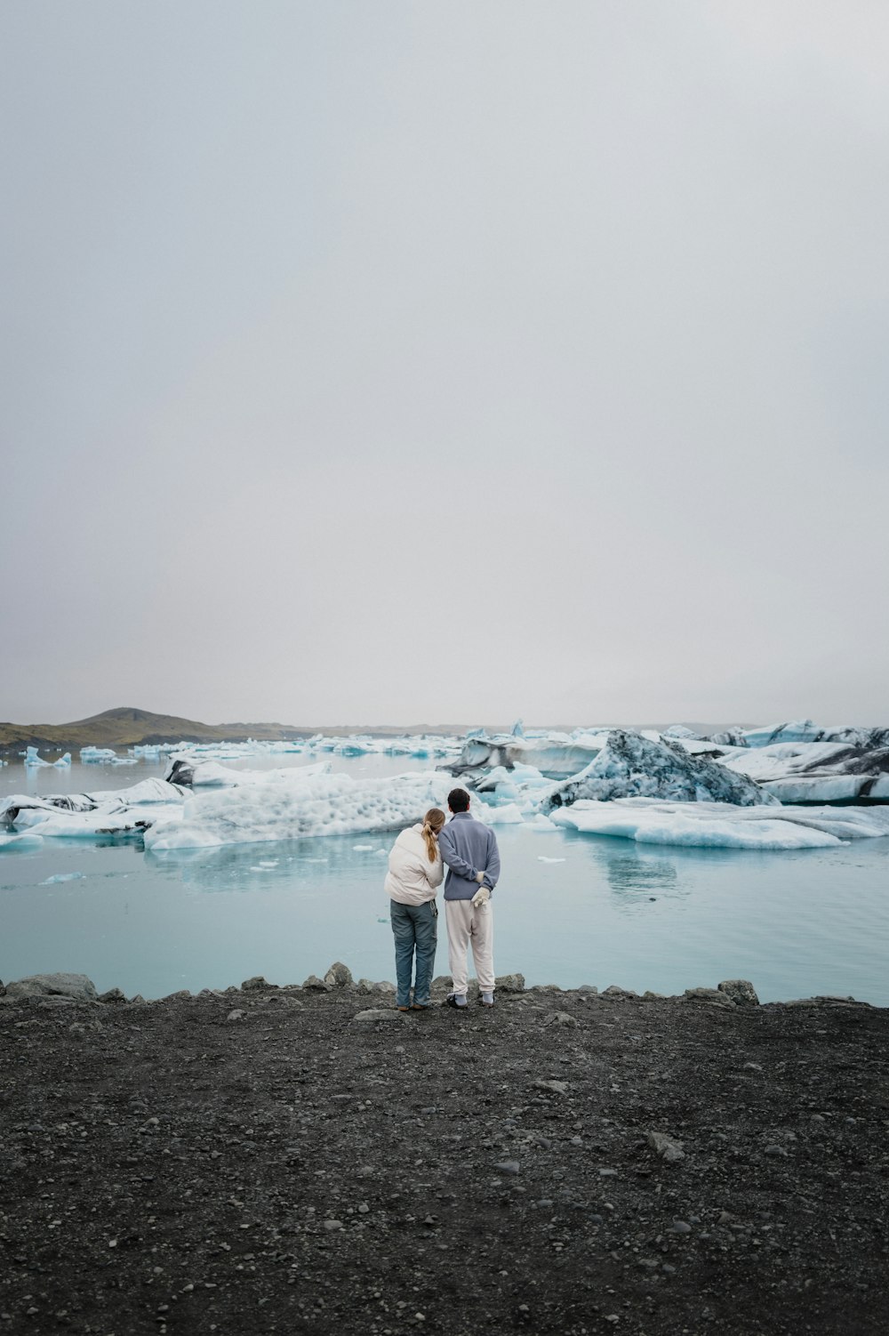 two people standing on a rocky beach next to a body of water