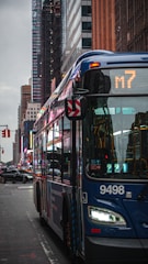 A city street scene featuring a blue public bus marked with route number M7. Skyscrapers and other buildings line the street in the background. The street is wet, possibly from rain, and other vehicles and some traffic signals are visible.