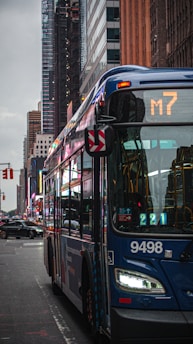 A city street scene featuring a blue public bus marked with route number M7. Skyscrapers and other buildings line the street in the background. The street is wet, possibly from rain, and other vehicles and some traffic signals are visible.