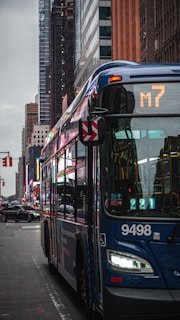 A city street scene featuring a blue public bus marked with route number M7. Skyscrapers and other buildings line the street in the background. The street is wet, possibly from rain, and other vehicles and some traffic signals are visible.