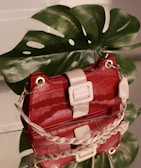 A glossy red handbag with a crocodile texture is placed on a reflective surface. The bag has beige accents, including a large rectangular buckle and braided handle. Behind the handbag is a large green monstera leaf, casting natural shadows.