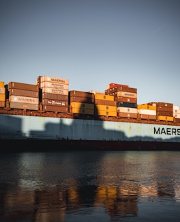 A large cargo ship carrying numerous containers is seen on calm waters. The containers are stacked in various colors, primarily yellow, brown, and red, and they bear the marking of the shipping company 'Maersk'. The photo is taken during a time of day when the sun casts long shadows, enhancing the rich colors of the containers and reflecting them on the surface of the water.