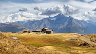 A serene mountain landscape with a cluster of rustic wooden cabins surrounded by vast meadows. Snow-capped peaks dominate the background under a dramatic sky filled with clouds. The scene is tranquil and expansive, with undulating hills leading up to majestic mountains.