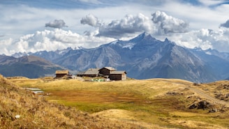 A serene mountain landscape with a cluster of rustic wooden cabins surrounded by vast meadows. Snow-capped peaks dominate the background under a dramatic sky filled with clouds. The scene is tranquil and expansive, with undulating hills leading up to majestic mountains.
