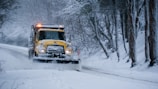 A yellow snowplow is clearing a snow-covered road surrounded by trees. The vehicle is pushing snow to the side as it drives through a winter landscape. The area is covered in heavy snow, and the trees have bare branches dusted with snow.