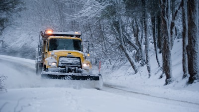 A yellow snowplow is clearing a snow-covered road surrounded by trees. The vehicle is pushing snow to the side as it drives through a winter landscape. The area is covered in heavy snow, and the trees have bare branches dusted with snow.