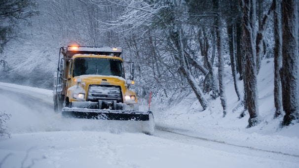 A yellow snowplow is clearing a snow-covered road surrounded by trees. The vehicle is pushing snow to the side as it drives through a winter landscape. The area is covered in heavy snow, and the trees have bare branches dusted with snow.