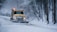 A yellow snowplow is clearing a snow-covered road surrounded by trees. The vehicle is pushing snow to the side as it drives through a winter landscape. The area is covered in heavy snow, and the trees have bare branches dusted with snow.