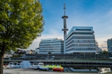 Modern office buildings with glass and concrete design stand beside a calm canal, reflecting the architectural structure. A tall communications tower rises prominently in the background. Several small boats dock near the edge of the water. Lush green trees frame the scene, and the sky is clear with a hint of twilight.