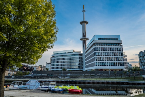 Modern office buildings with glass and concrete design stand beside a calm canal, reflecting the architectural structure. A tall communications tower rises prominently in the background. Several small boats dock near the edge of the water. Lush green trees frame the scene, and the sky is clear with a hint of twilight.