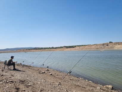 A person sitting on a foldable chair by the side of a calm riverbank holding a fishing rod. Several other fishing rods are planted in the ground along the rocky shore. The expansive blue sky stretches over the tranquil, sparsely vegetated landscape with a distant shoreline and hills.