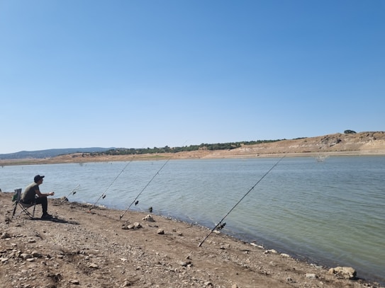A person sitting on a foldable chair by the side of a calm riverbank holding a fishing rod. Several other fishing rods are planted in the ground along the rocky shore. The expansive blue sky stretches over the tranquil, sparsely vegetated landscape with a distant shoreline and hills.
