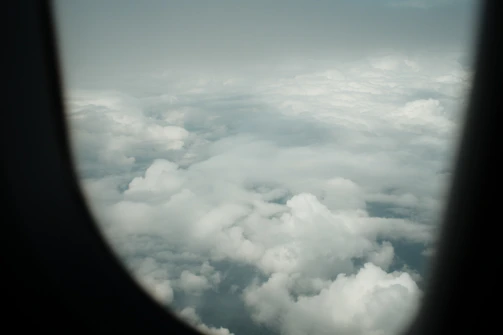 A cozy neck pillow draped over a plane seat headrest with a window view of clouds outside.