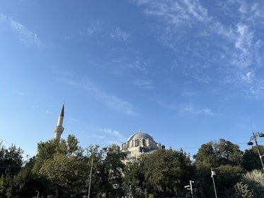 A mosque with a prominent dome and a tall minaret surrounded by lush green trees. The sky is clear with scattered clouds, and the lighting suggests a bright, sunny day.
