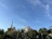 A mosque with a prominent dome and a tall minaret surrounded by lush green trees. The sky is clear with scattered clouds, and the lighting suggests a bright, sunny day.