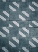 A pattern of dark gray tiles with white diagonal stripes, arranged in a grid. The surface appears rough and textured, giving an industrial or urban feel.