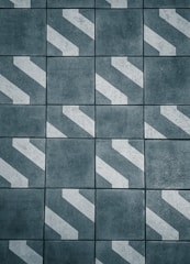 A pattern of dark gray tiles with white diagonal stripes, arranged in a grid. The surface appears rough and textured, giving an industrial or urban feel.
