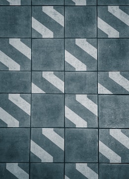 A pattern of dark gray tiles with white diagonal stripes, arranged in a grid. The surface appears rough and textured, giving an industrial or urban feel.