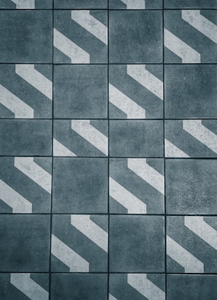 A pattern of dark gray tiles with white diagonal stripes, arranged in a grid. The surface appears rough and textured, giving an industrial or urban feel.