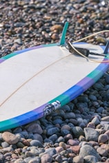 A surfboard with colorful edges lies on a bed of smooth pebbles. The board is primarily white with a blue, purple, and green border, and a leash is attached to it.