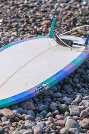A surfboard with colorful edges lies on a bed of smooth pebbles. The board is primarily white with a blue, purple, and green border, and a leash is attached to it.