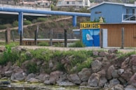 A rustic scene featuring a small, painted building with the sign 'BAJAMIGOS'. The building is partially enclosed by a chain-link fence and surrounded by green vegetation. Rocks and greenery are visible in the foreground, and a large blue pipeline stretches across the background. There are residential buildings in the distance.