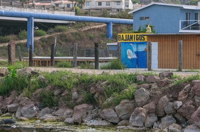 A rustic scene featuring a small, painted building with the sign 'BAJAMIGOS'. The building is partially enclosed by a chain-link fence and surrounded by green vegetation. Rocks and greenery are visible in the foreground, and a large blue pipeline stretches across the background. There are residential buildings in the distance.