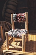 A small, handcrafted wooden chair with a woven seat and backrest featuring multicolored threads. The chair is placed on a wicker surface, and the scene is illuminated by natural light, casting shadows and adding warmth to the overall composition.