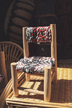 A small, handcrafted wooden chair with a woven seat and backrest featuring multicolored threads. The chair is placed on a wicker surface, and the scene is illuminated by natural light, casting shadows and adding warmth to the overall composition.