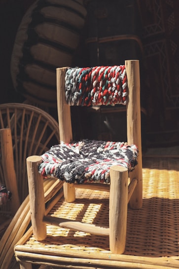 A small, handcrafted wooden chair with a woven seat and backrest featuring multicolored threads. The chair is placed on a wicker surface, and the scene is illuminated by natural light, casting shadows and adding warmth to the overall composition.