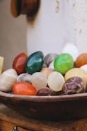 A collection of polished, colorful stones in various shapes and sizes displayed in a wooden bowl. The stones include vibrant shades of green, red, yellow, and white, and some have a marbled texture. The background appears to be a light-colored wall, adding to the rustic and natural setting.