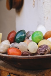A collection of polished, colorful stones in various shapes and sizes displayed in a wooden bowl. The stones include vibrant shades of green, red, yellow, and white, and some have a marbled texture. The background appears to be a light-colored wall, adding to the rustic and natural setting.