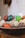 A collection of polished, colorful stones in various shapes and sizes displayed in a wooden bowl. The stones include vibrant shades of green, red, yellow, and white, and some have a marbled texture. The background appears to be a light-colored wall, adding to the rustic and natural setting.