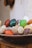 A collection of polished, colorful stones in various shapes and sizes displayed in a wooden bowl. The stones include vibrant shades of green, red, yellow, and white, and some have a marbled texture. The background appears to be a light-colored wall, adding to the rustic and natural setting.