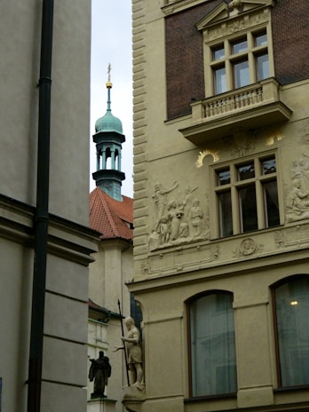 A decorative stone facade on a historic building features intricate carvings and sculptures. The building includes ornate window frames and detailed reliefs. In the background, a copper-green spire with a cross tops a red-tiled roof, set against a cloudy sky.