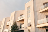 A modern residential building with multiple stories and large glass windows, featuring a sleek, minimalist design. The exterior is composed of vertical metal panels, and there are glass balconies. The sunlight casts a warm glow over the facade, and a small tree stands in front of the building.