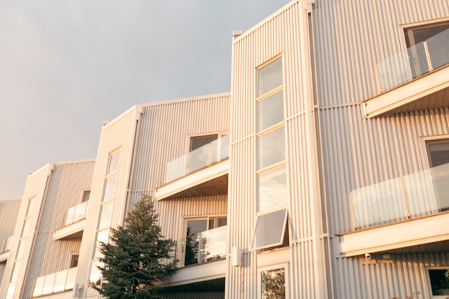 A modern residential building with multiple stories and large glass windows, featuring a sleek, minimalist design. The exterior is composed of vertical metal panels, and there are glass balconies. The sunlight casts a warm glow over the facade, and a small tree stands in front of the building.