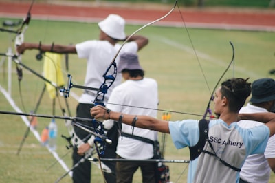 Several archers are standing on a grassy field, each aiming their bows and arrows towards a target that is out of sight. They are equipped with professional archery gear, including arm guards and quivers.
